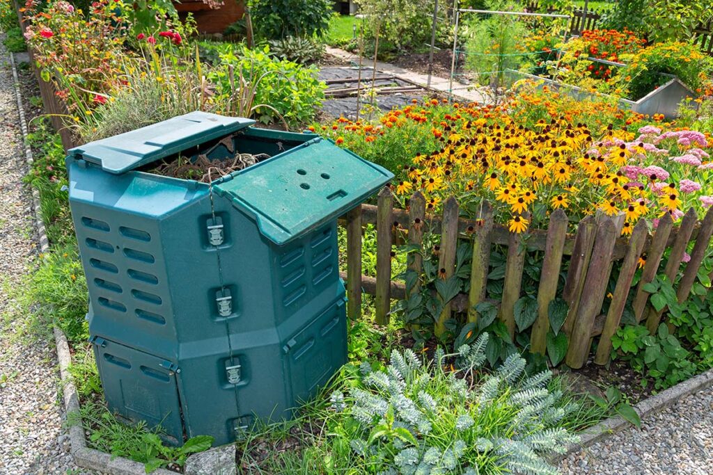 Kompostieren easy: Ein geöffneter Thermo-Komposter vor prächtig blühenden Blumen im Familiengarten Areal Buen, Ende Sommer, sozusagen eine Gratis-Kompostabgabe